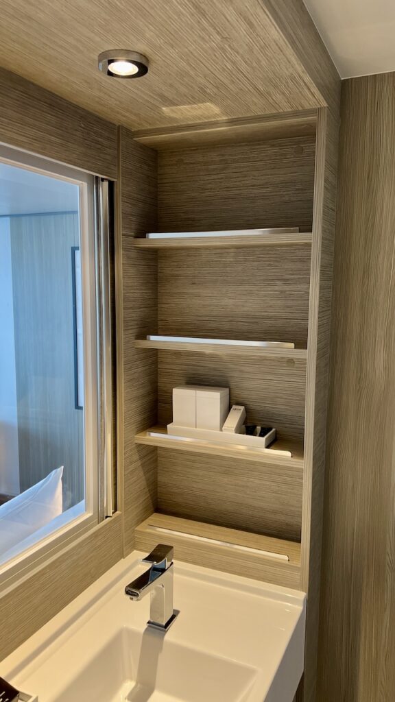 View of storage at the right side of the sink, with four open shelves. One holds complimentary toiletry amenities.