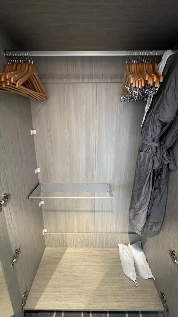 Spacious and well-organized closet spaces in Celebrity Ascent Sky Suite, offering ample storage for clothing and personal items. On the left, a closet with one long rod, a shorter rod beneath, wood clothes hangers, two bathrobes, and complimentary slippers.