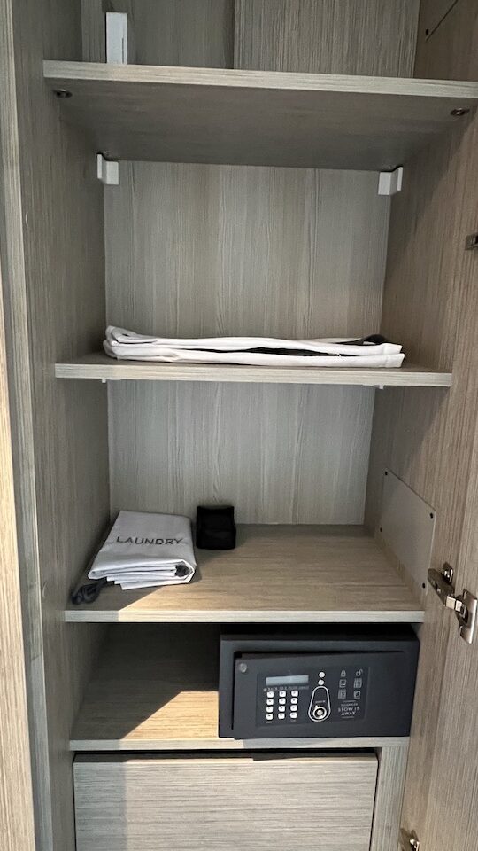 The second closet in the Celebrity Ascent Sky Suite, which has four open shelves. On the shelves are a complimentary tote bag, a laundry bag, compact binoculars for your use, and a mini safe. Below are three drawers.