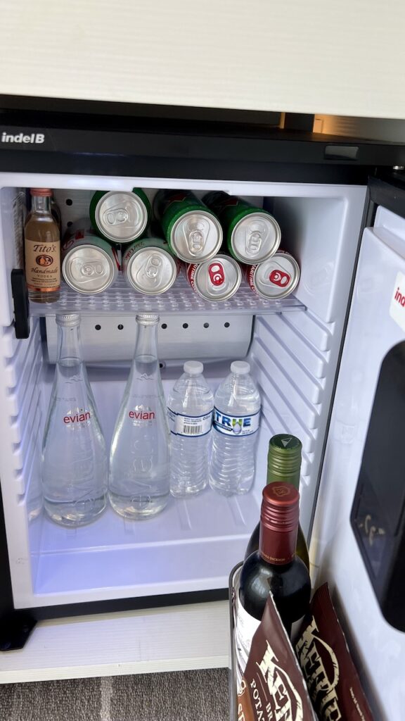 Interior of the mini fridge in the Celebrity Sky Suite, holding cans of soda, bottles of water, bottles of wine, chips, and mini bottles of liquor. These items are for sale.