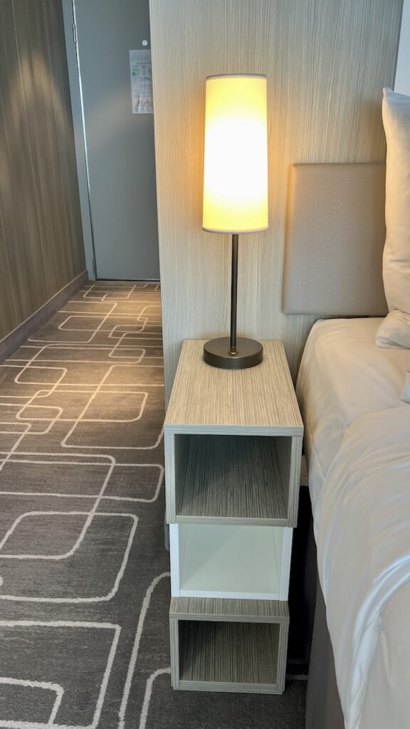 Left nightstand of the Sky Suite, narrow with three cubbies and a tall, slim light affixed to its surface. No electrical outlets.