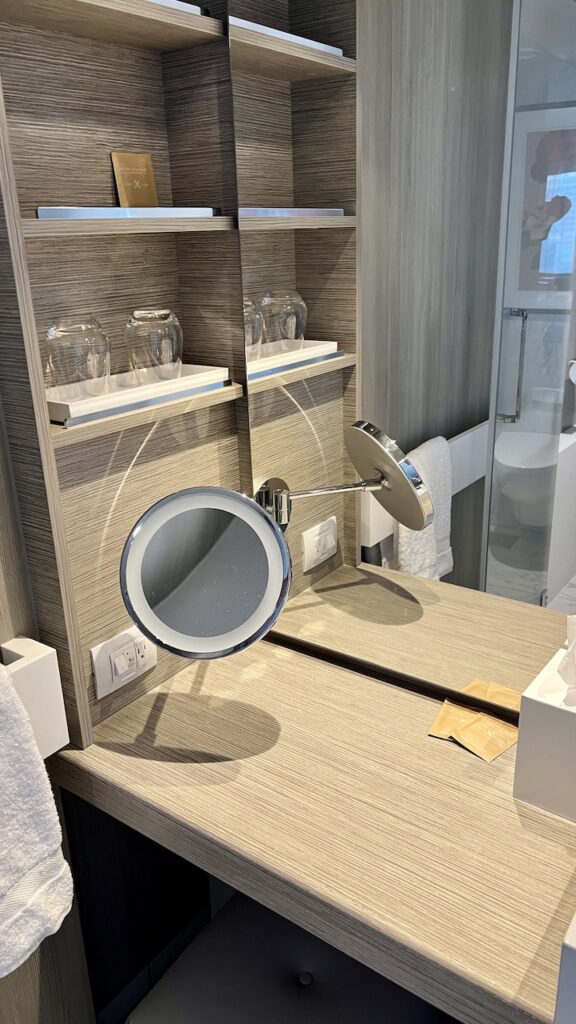 Makeup area consisting of a small desk with a hassock beneath. Affixed to the wall is a lighted magnifying mirror. Storage on the wall features four open shelves, one holding two glasses. A US electrical outlet is on the wall.